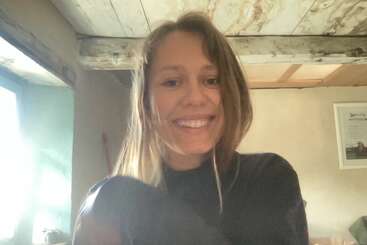 A young woman with long hair smiles warmly at the camera in a cozy, rustic room with wooden ceilings and soft daylight filling the space.