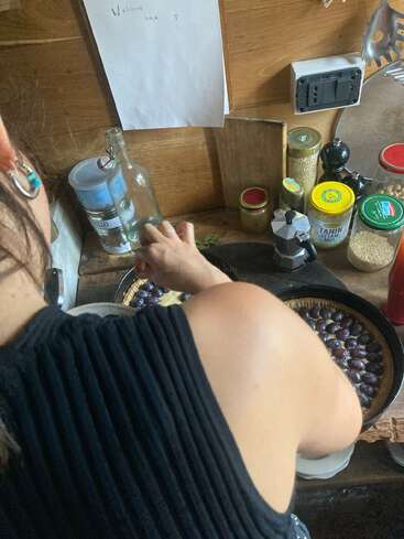 Eine Person arrangiert Trauben auf einer Torte in einer gemütlichen Küche. In der Nähe stehen verschiedene Gläser, Gewürze und Utensilien. Darüber hängt ein handgeschriebener "Willkommen zurück"-Zettel.