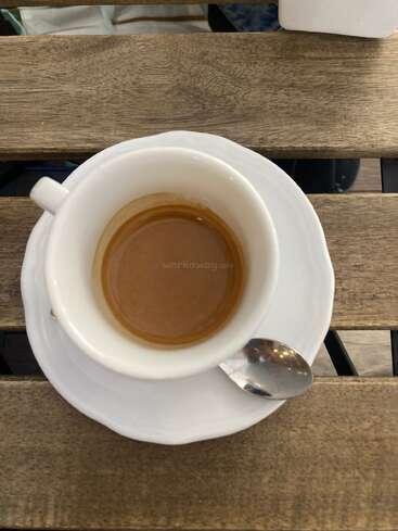 Eine weiße Tasse mit Espresso steht auf einer passenden Untertasse auf einem Holztisch. Ein kleiner silberner Löffel ruht auf der Untertasse und schafft ein gemütliches Ambiente.