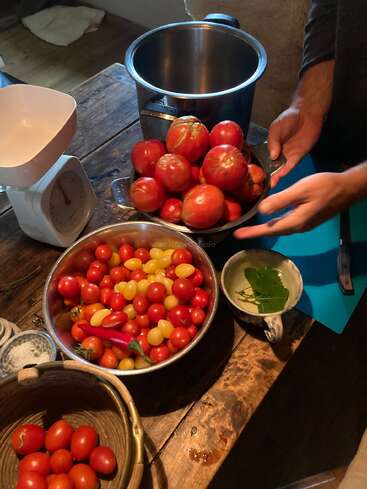 Eine rustikale Küchenszene zeigt verschiedene Schüsseln mit frischen Tomaten, eine Küchenwaage, einen Topf, Kräuter in einer Tasse und Hände, die Zutaten auf einem Holztisch zubereiten.