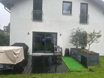 A modern white house with three upper windows, sliding glass doors, small balcony railings, a wet wooden deck, covered furniture, planters, barbecue, and lush green lawn.