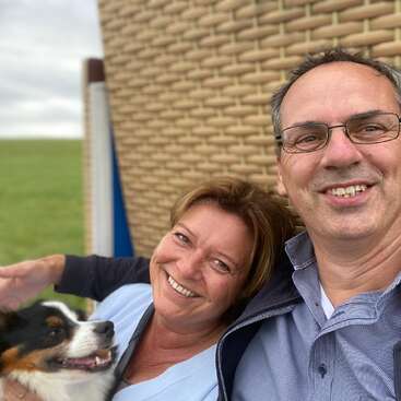 A smiling couple takes a selfie outdoors, sitting near a wicker structure. The woman holds a happy dog, and green grass is visible in the background.