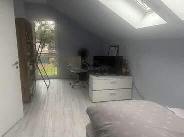 A cozy, light-filled bedroom with slanted ceiling, skylight, and white decor. Features a bed, TV, drawers, bookshelf, desk, chair, and garden view.