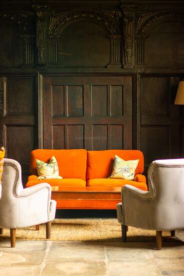 Ein orangefarbenes Sofa mit gemusterten Kissen steht zwei hellen Sesseln gegenüber. Verschnörkelte dunkle Holzvertäfelungen und eine hohe Lampe schaffen ein elegantes, altes und gemütliches Ambiente.