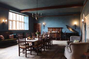 Dieser Raum hat blaue Wände, einen langen Esstisch aus Holz mit Stühlen, einen Kronleuchter, einen Flügel, gemütliche Sessel, große Fenster und eine dekorative Beleuchtung.