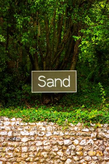 Eine Steinmauer grenzt an üppiges grünes Laub und dichte Bäume. Davor steht ein Schild mit der humorvollen Aufschrift "Sand", obwohl nirgendwo Sand zu sehen ist.