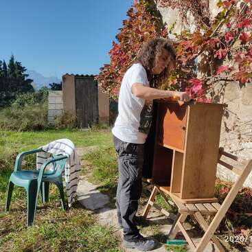 Eine Person mit langen Haaren steht draußen und schleift einen Holzschrank. Ein Stuhl und ein Handtuch stehen daneben, umgeben von einem Garten und einem klaren blauen Himmel.