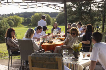 A group of people enjoy an outdoor meal together under a canopy with string lights, surrounded by greenery and sunshine, creating a warm and cheerful atmosphere.