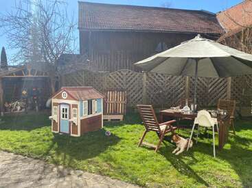 Un patio trasero soleado cuenta con una casita de juegos, sillas de madera, una mesa de comedor con vajilla, una trona, una sombrilla, un perro descansando y un césped verde y frondoso.