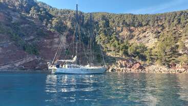 Un voilier blanc flotte paisiblement sur une eau calme et limpide près d'une côte rocheuse et boisée, sous un ciel bleu éclatant, entouré d'arbres verdoyants et de falaises.