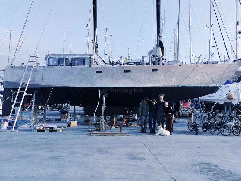 Un grand voilier est posé sur des supports dans une marina. Quatre personnes et un chien posent en dessous. Des échelles, des vélos et des outils les entourent, et d'autres bateaux se trouvent à proximité.