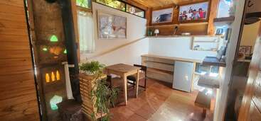 Ein gemütlicher Holzraum mit einem kleinen Tisch, zwei Stühlen, einem Minikühlschrank, einem Holzofen, Zimmerpflanzen, einem Regal, Wandkunst und warmem, natürlichem Sonnenlicht.