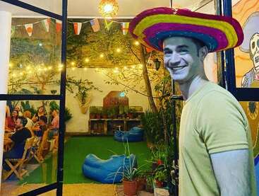 A smiling man wearing a colorful sombrero stands at a doorway, while people gather and chat inside a festive, warmly-lit, decorated patio with flags.