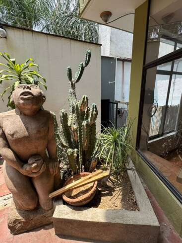 A stone statue kneels next to a potted cactus and plant in a sunny courtyard, bordered by beige walls, reflecting windows, and a tiled floor.
