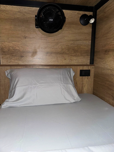 A neatly made single bed with a white pillow and sheet, wooden walls, a small black fan, spotlight, and an electrical socket on the headboard.