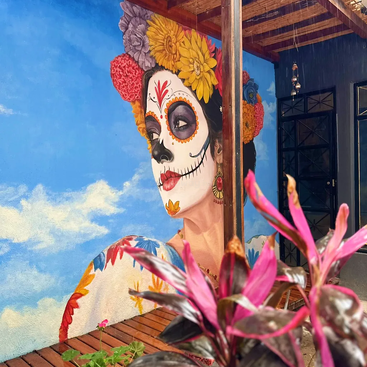 A vibrant mural depicts a woman with Día de los Muertos face paint and colorful flowers in her hair. Pink plants and blue sky add contrast.