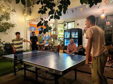 Four young men enjoy a nighttime game of ping pong indoors, surrounded by colorful wall art, hanging lights, and a relaxed, social atmosphere in a cozy café.