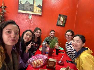 Seis amigos estão sentados em torno de uma mesa vermelha num restaurante aconchegante com paredes vermelhas brilhantes, sorrindo e curtindo uma refeição juntos, rodeados por uma decoração colorida.