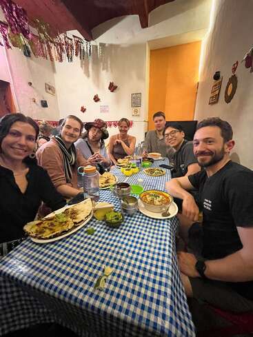 Um grupo de sete amigos está curtindo uma refeição juntos em um restaurante aconchegante com decoração festiva. Todos parecem felizes, rodeados por uma deliciosa comida mexicana.