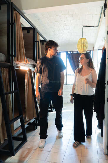 Um homem e uma mulher estão conversando em um quarto bem iluminado de albergue, com beliches, cortinas bege e um abajur de tecido no teto. A luz do sol entra pelas janelas.
