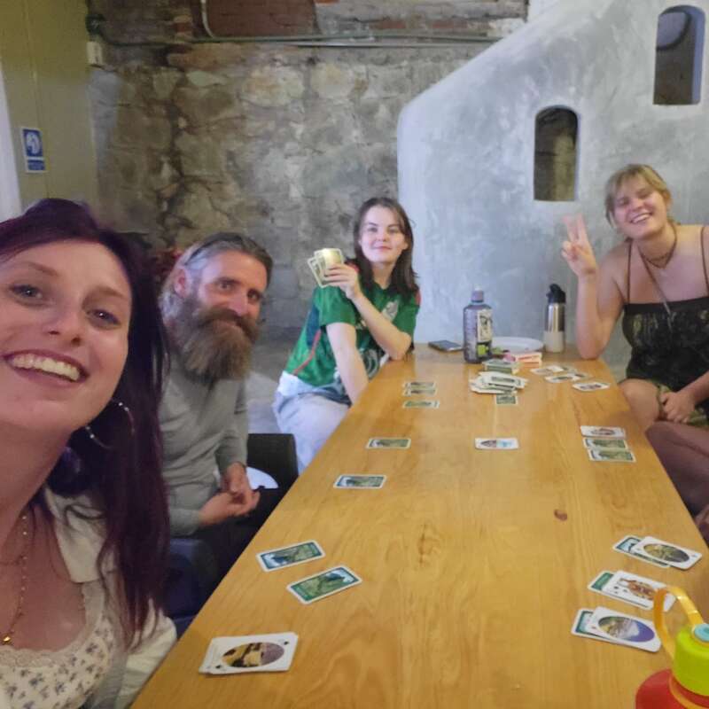 Quatro amigos estão sentados ao redor de uma mesa de madeira, sorrindo e posando enquanto jogam cartas. O ambiente é rústico, com paredes de pedra e um clima descontraído e alegre.