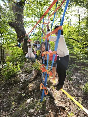 Several children are balancing and playing on a colorful rope course in a lush, green forest, surrounded by trees, sunlight, and natural elements, enjoying outdoor adventure.