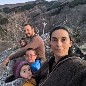 Uma família de cinco pessoas está sentada no topo de uma colina rochosa, cercada por montanhas e árvores sem folhas, curtindo a natureza juntos. Todos parecem calmos e felizes.
