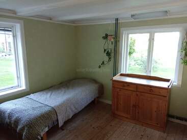 Das ist ein schlichtes, gemütliches Schlafzimmer mit einem Einzelbett, grün gestrichenen Wänden, einem Holzboden, einer Kommode, zwei Fenstern und ein paar Hängepflanzen.