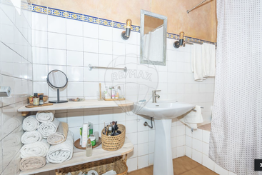 This image shows a tidy bathroom with white tiles, a pedestal sink, shelves holding rolled towels, toiletries, a mirror, and a shower with a curtain.