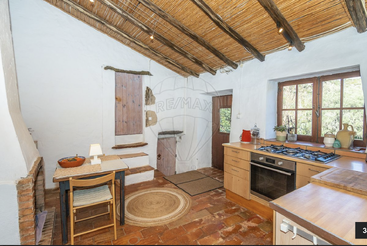 This cozy rustic kitchen features a wooden beamed ceiling, terracotta floor, whitewashed walls, small dining area, modern stove, and large windows offering beautiful natural light.