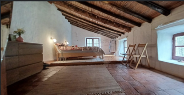 A cozy attic bedroom with rustic wooden beams, white plaster walls, and terracotta tile flooring. The bed sits near two windows; minimalistic, warm, inviting charm.