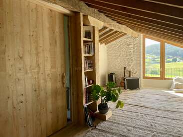 Esta acogedora habitación abuhardillada cuenta con vigas de madera, una puerta corredera de madera, una estantería, una maceta, una gran ventana con vistas panorámicas, instrumentos musicales y luz natural que llena el espacio.