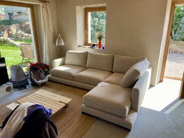 Una sala de estar iluminada por el sol, con un sofá seccional beige, una mesa de centro de madera, ropa esparcida, enseres de bebé y grandes ventanales con vistas a un verde patio trasero.