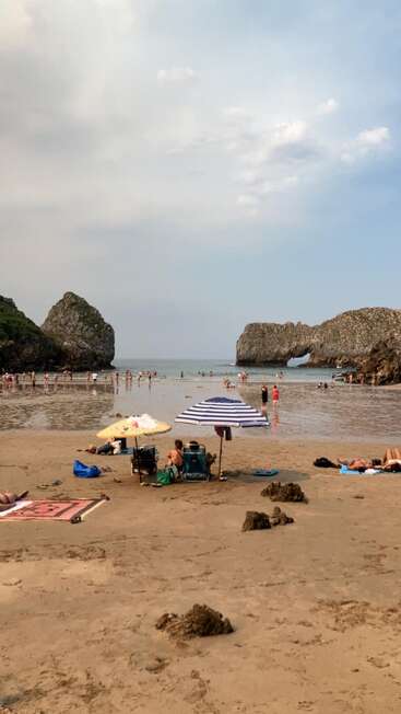 La gente se relaja en una playa de arena, algunos bajo sombrillas. Otros juegan en aguas poco profundas cerca de acantilados rocosos. El cielo está parcialmente nublado. Una escena tranquila y veraniega.