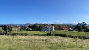 Esta imagen muestra un pintoresco paisaje rural, con campos verdes cubiertos de hierba, casas dispersas, montañas lejanas, cielo azul y un ambiente rural tranquilo y apacible bañado por la luz del sol.