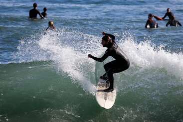 Un surfista cabalga hábilmente una ola, creando un chorro de agua, mientras varios otros surfistas con trajes de neopreno flotan en el fondo, esperando su turno.