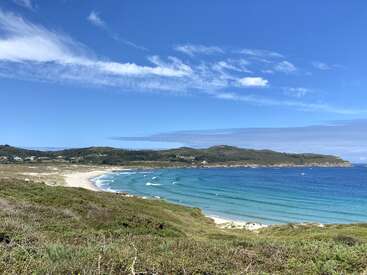 Un cielo azul despejado se extiende sobre un hermoso paisaje costero, con olas turquesas que se encuentran suavemente con una playa de arena. Las colinas ondulantes y la escasa vegetación completan esta vista tranquila y pintoresca.