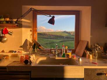 A sunlit kitchen sink area with dishes, utensils, and cleaning supplies. Through the window, green fields and a blue sky with clouds are beautifully visible.