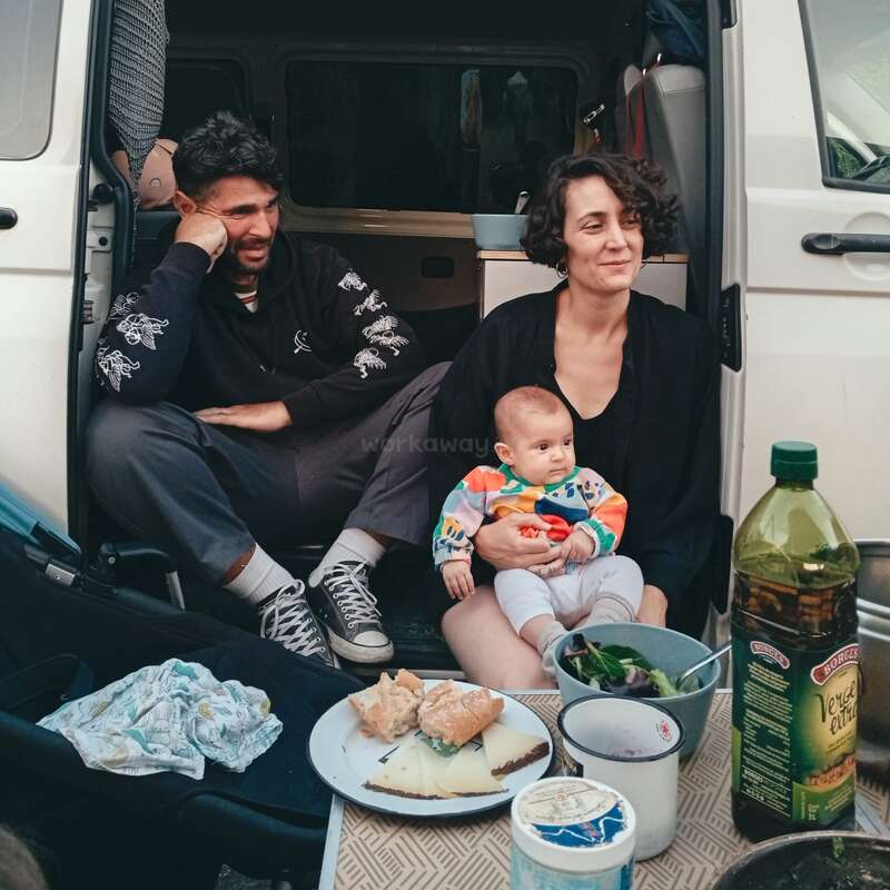 Una familia sentada a la puerta abierta de una furgoneta, disfrutando de una comida informal al aire libre. La madre sostiene a un bebé; se ven alimentos y aceite de oliva.