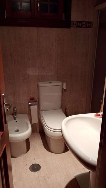 This image shows a small bathroom with a toilet, bidet, and sink. Beige tiles cover the walls and floor. A wooden window is above.