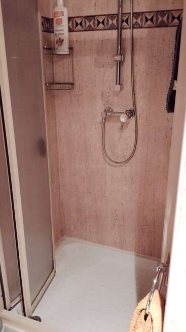 This image shows a small shower with beige tiled walls, a sliding glass door, metal shower head, soap rack, shampoo bottle, and a tan towel hanging nearby.