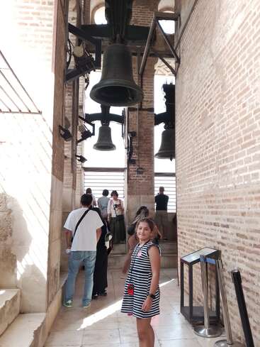 A girl in a striped dress stands near large historic bells in a tower. Other visitors are observing and taking in the brick architecture and sunlight.