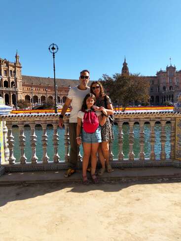 A family of three poses on a sunny day by a decorative railing, with a historic building, canal, and clear blue sky in the background.