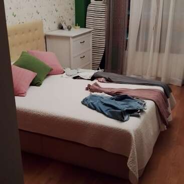 A cozy bedroom with a double bed, colorful pillows, and scattered clothes. There’s a white dresser, ironing board, sheer curtains, and wooden flooring in the room.