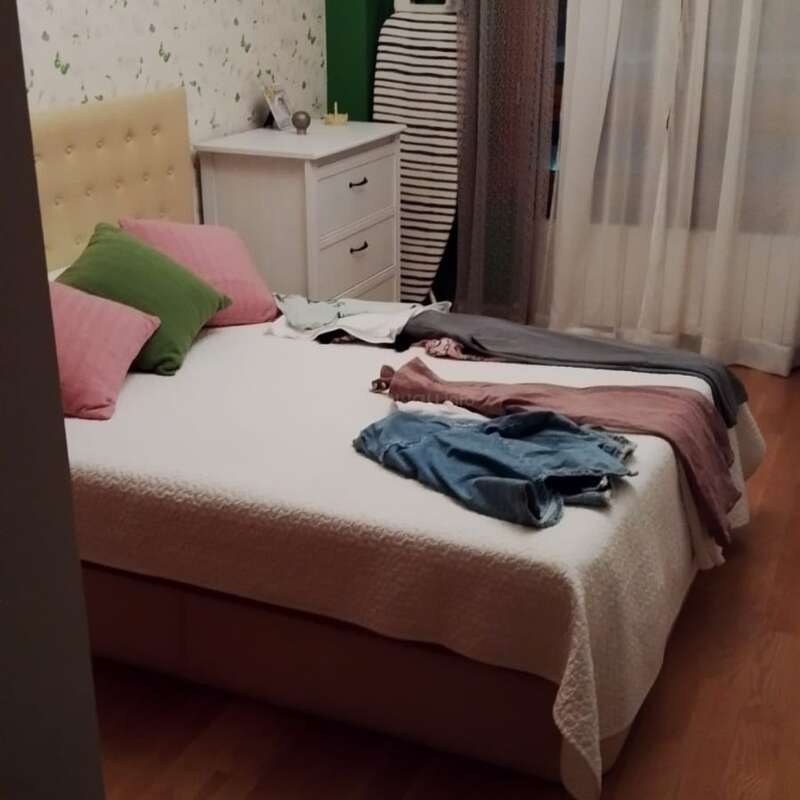 A cozy bedroom with a double bed, colorful pillows, and scattered clothes. There’s a white dresser, ironing board, sheer curtains, and wooden flooring in the room.