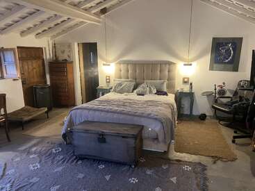 Este quarto aconchegante apresenta uma cama grande com cabeceira tufada, móveis rústicos, baú de madeira, tapetes, iluminação ambiente, cadeira de escritório e teto com vigas expostas.