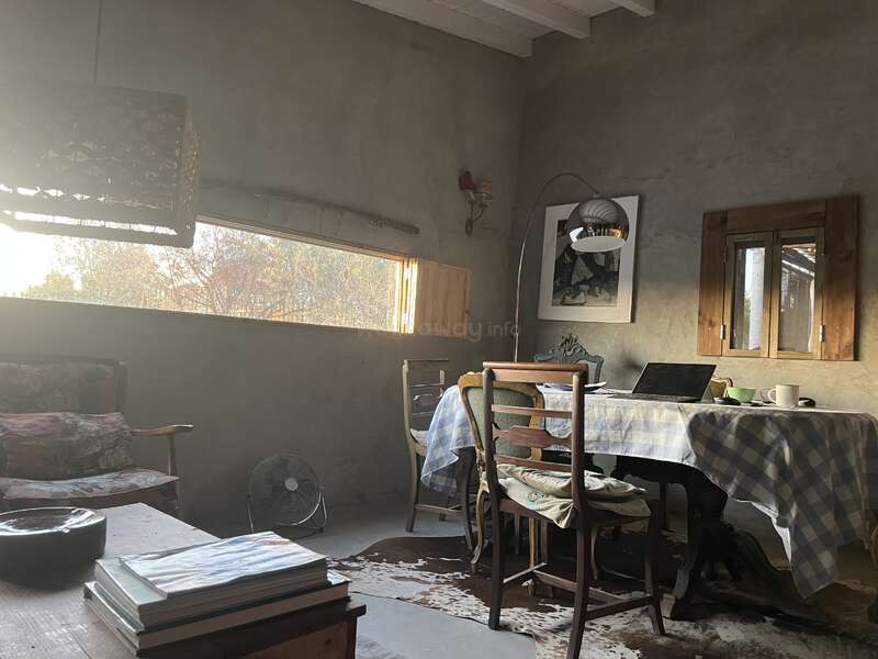 A luz do sol entra em uma sala rústica com uma configuração simples: cadeiras de madeira, mesa com tecido xadrez, livros, um sofá e uma janela com persianas de madeira. Ambiente aconchegante.