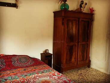 Une chambre chaleureuse avec un couvre-lit aux motifs colorés, une petite table de chevet en bois et une grande armoire vintage en bois surmontée d'objets décoratifs et d'un vase vert.
