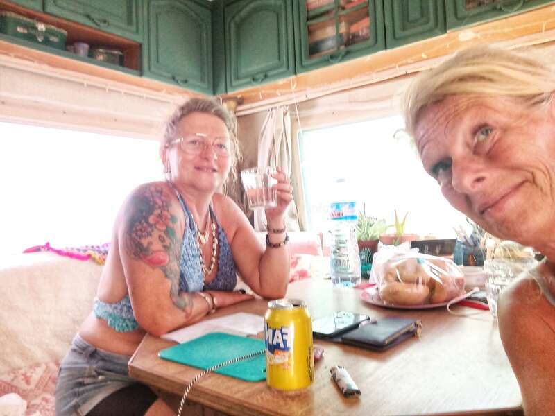 Deux femmes se détendent dans un camping-car confortable : l'une sourit à la caméra, l'autre tient un verre à la main. De la nourriture, des boissons et des objets du quotidien sont disposés sur la table.