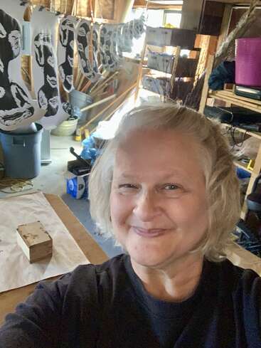Eine lächelnde Frau macht ein Selfie in einer Werkstatt. Im Hintergrund trocknen auf einer Wäscheleine im Siebdruckverfahren bedruckte Socken mit schwarzen und weißen Motiven.